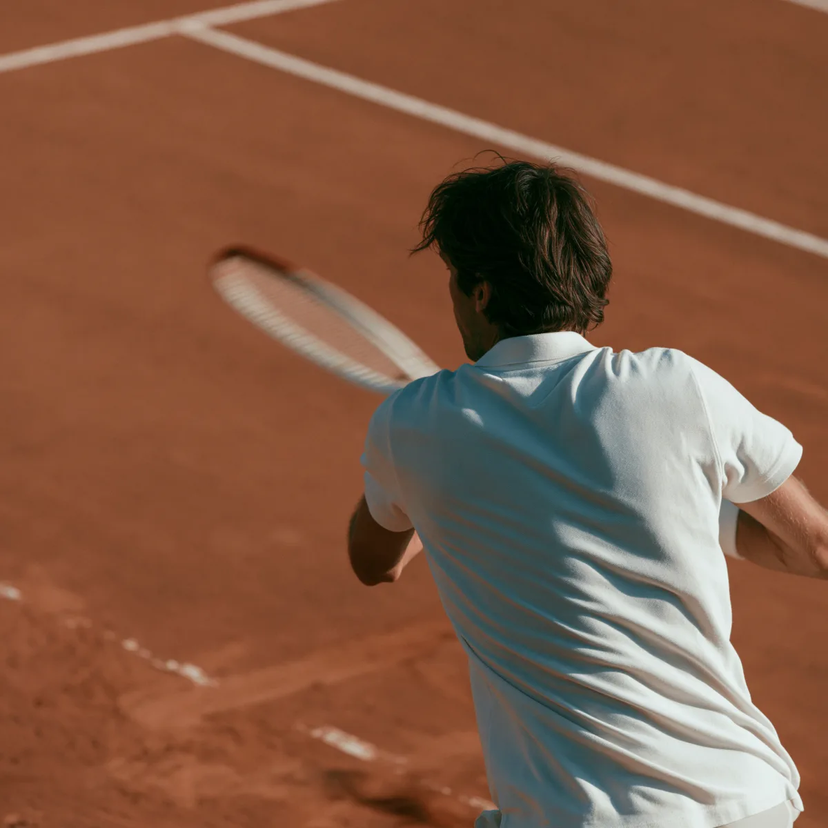 Person im weißen Polo spielt Tennis auf einem roten Sandplatz, von hinten